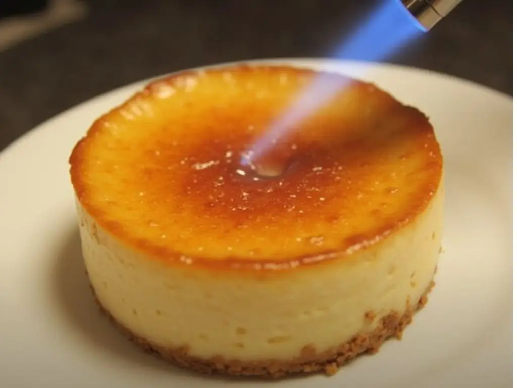 A kitchen torch caramelizing the sugar topping on a Crème Brûlée Cheesecake, creating a glossy golden-brown crust.