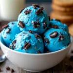 A bowl of bright blue Cookie Monster ice cream with chunks of Oreo and chocolate chip cookies, served on a rustic wooden table.