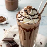 A creamy chocolate milkshake in a tall glass topped with whipped cream, chocolate syrup, and chocolate shavings, served with a straw.