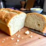 A sliced loaf of homemade gluten-free bread with a golden crust and soft, airy texture on a wooden cutting board, accompanied by a dish of butter.