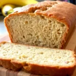 Freshly baked 4 ingredient banana bread with a golden crust and soft, moist inside, sliced on a wooden cutting board with ripe bananas in the background.