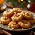 A plate of golden-brown Greek melomakarona cookies topped with walnuts and drizzled with honey syrup, served with cinnamon sticks and orange slices.